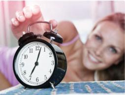 Imagem de um despertador com uma mulher em segundo plano a esticar-se para o desligar.